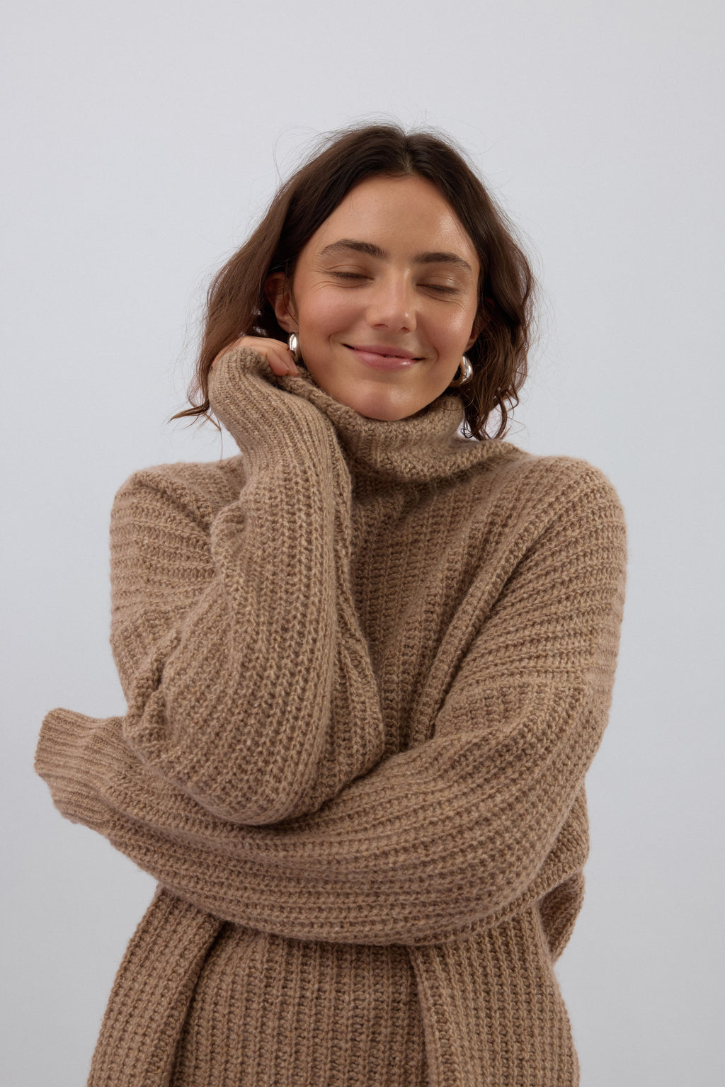Woman wearing a brown knitted sweater against a white background with silver earrings