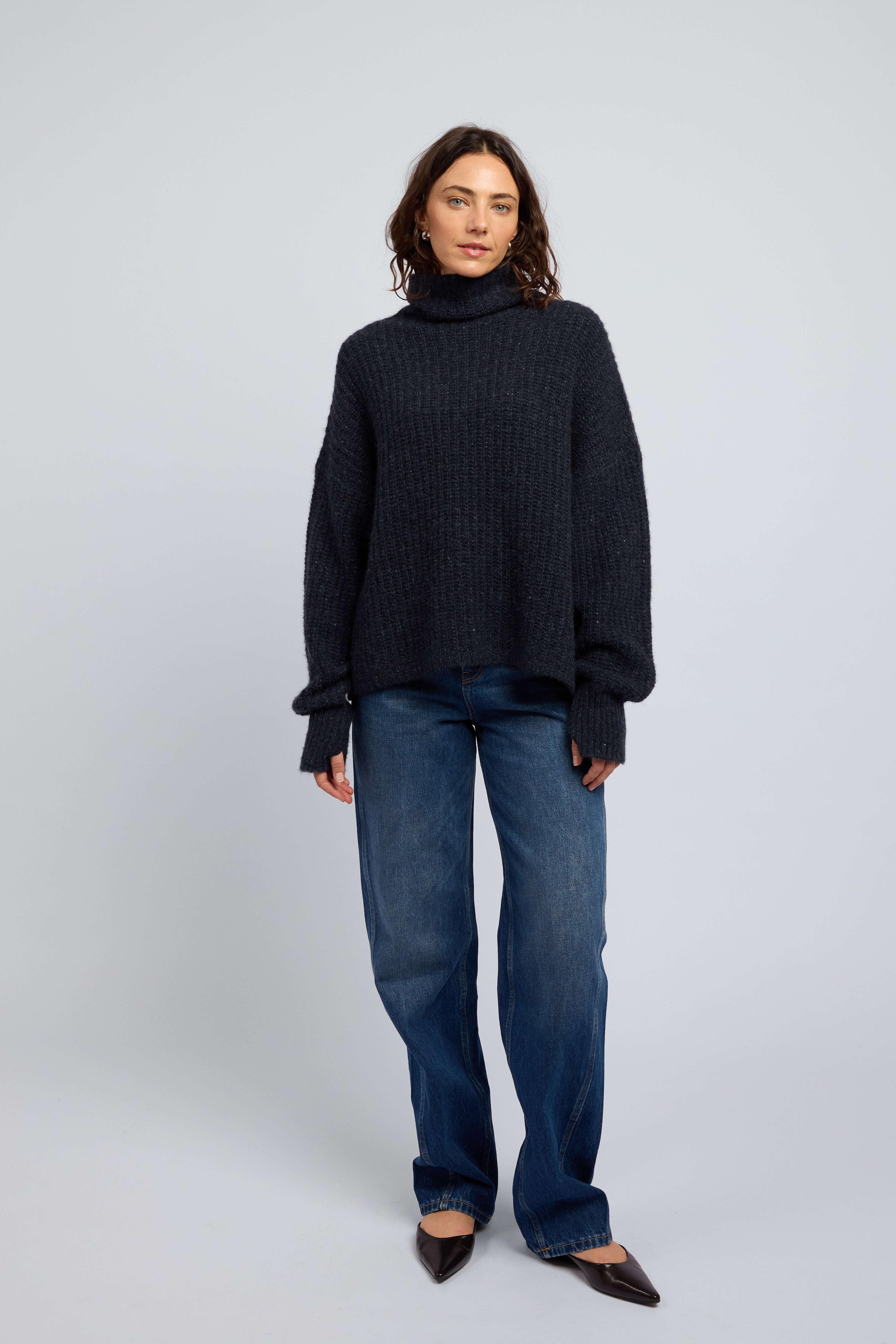 Woman wearing a dark midnight navy cashmere turtleneck sweater and blue jeans on a white background with black point toe flats