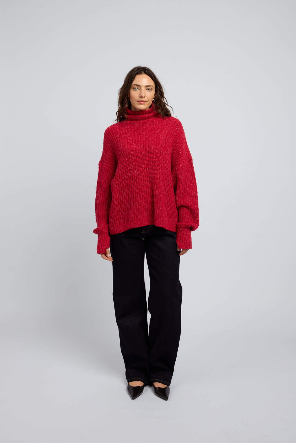 Woman wearing a poppy red cashmere turtleneck sweater and blue jeans on a white background with black point toe flats