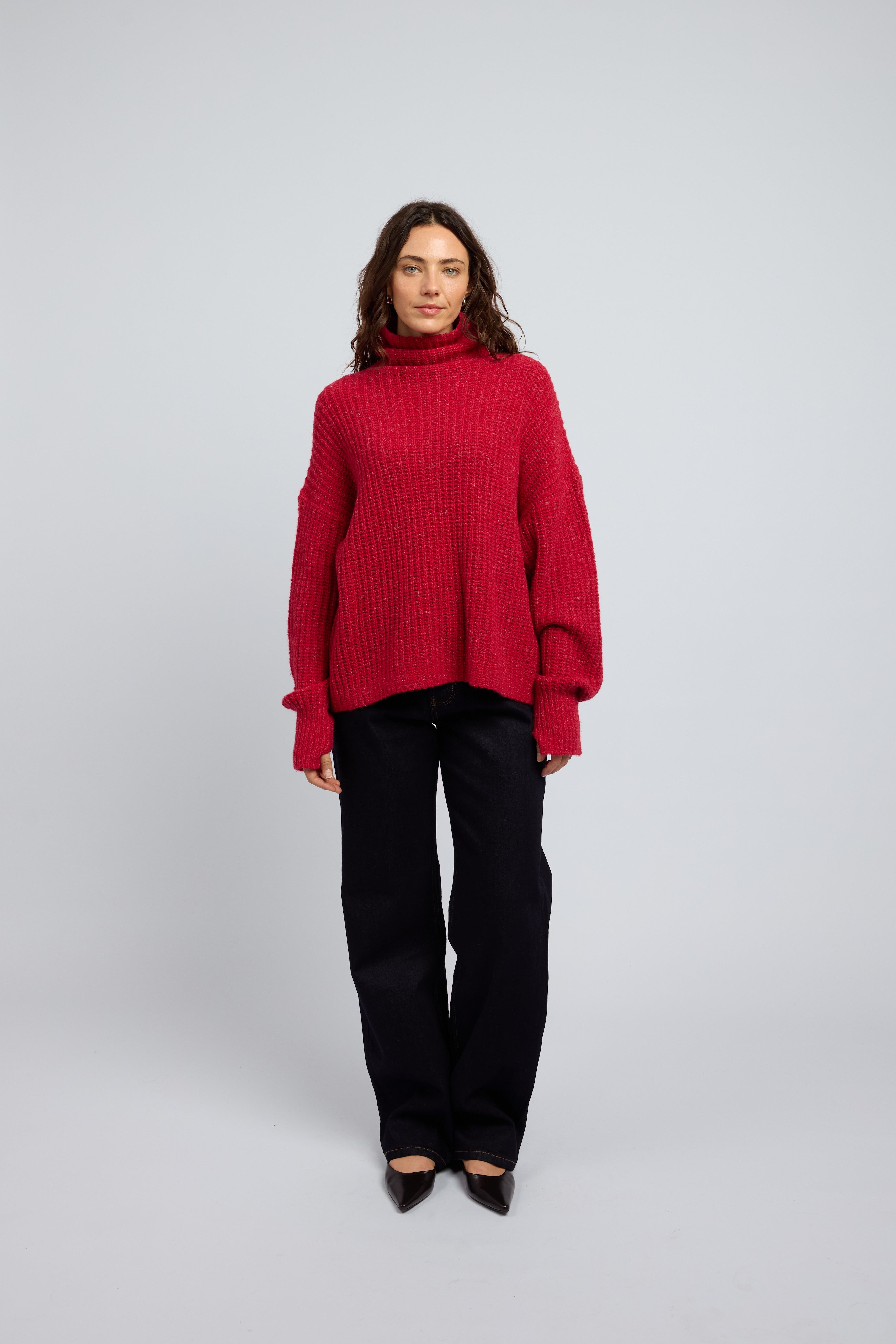 Woman wearing a poppy red cashmere turtleneck sweater and blue jeans on a white background with black point toe flats