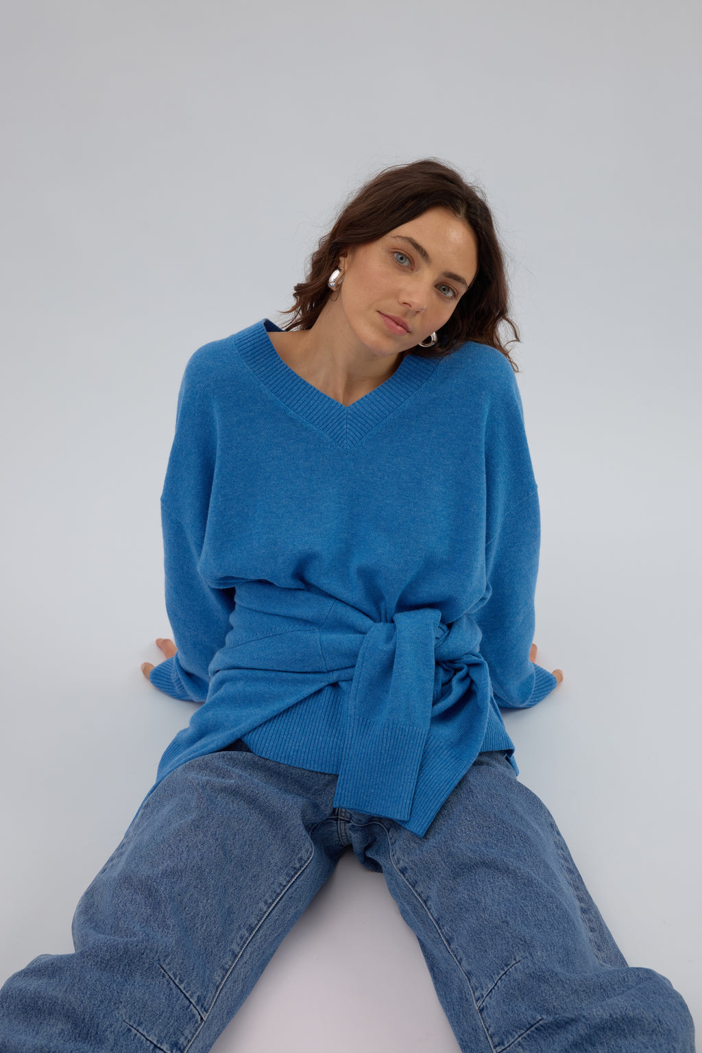 Woman sitting down with a bold blue v neckcashmere long sleeved sweater and white blue jeans, another knit is wrapped around her waist for extra styling
