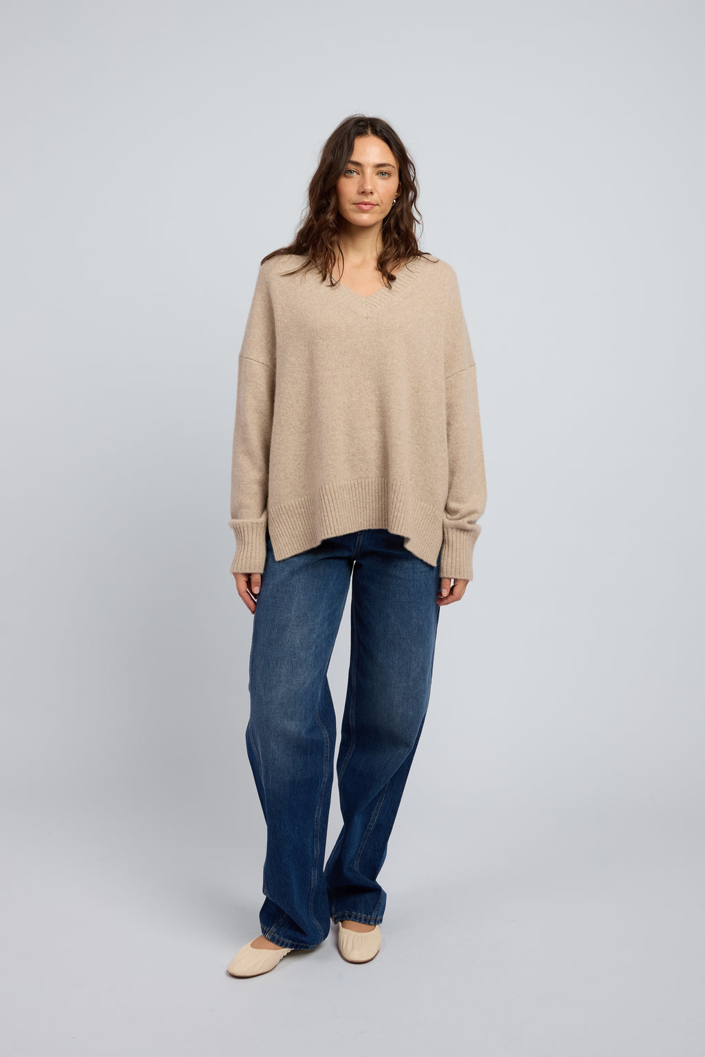 Woman wearing a slouchy beige cashmere long sleeve jumper in Oat with ribbed cuffs and neckline, paired with blue jeans and cream ballet flats