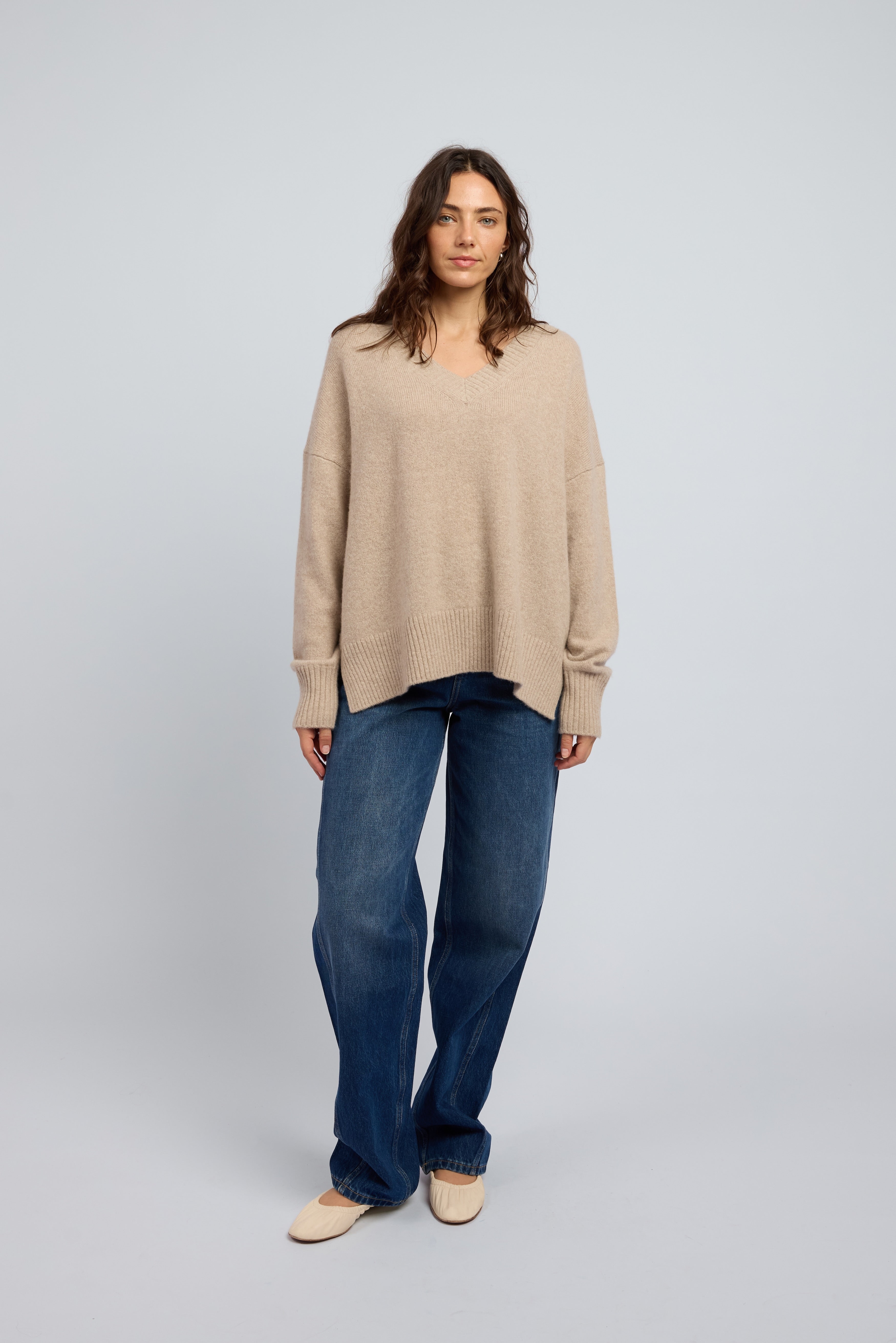 Woman wearing a slouchy beige cashmere long sleeve jumper in Oat with ribbed cuffs and neckline, paired with blue jeans and cream ballet flats