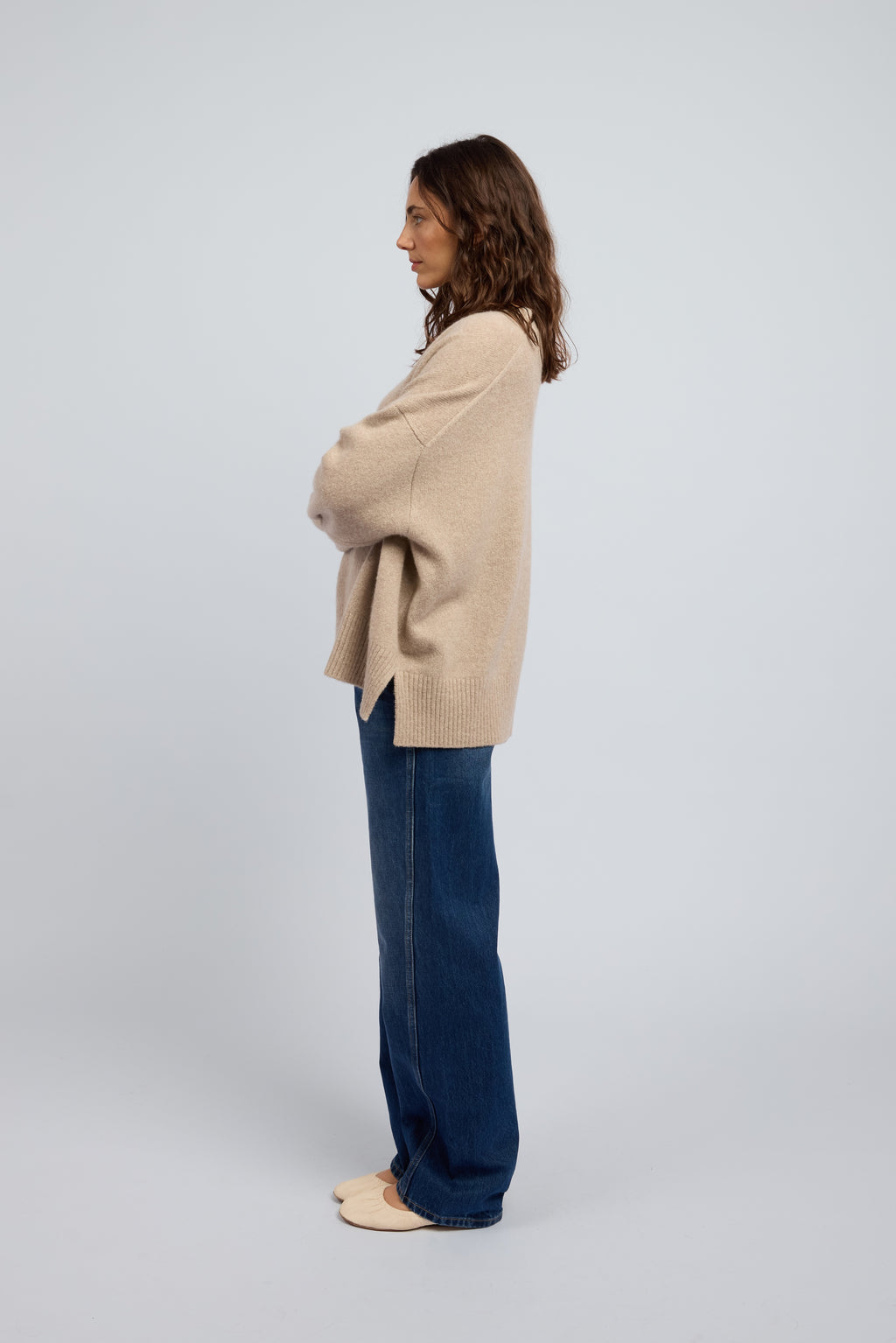 Woman wearing a slouchy beige cashmere long sleeve jumper in Oat with ribbed cuffs and neckline, paired with blue jeans and cream ballet flats