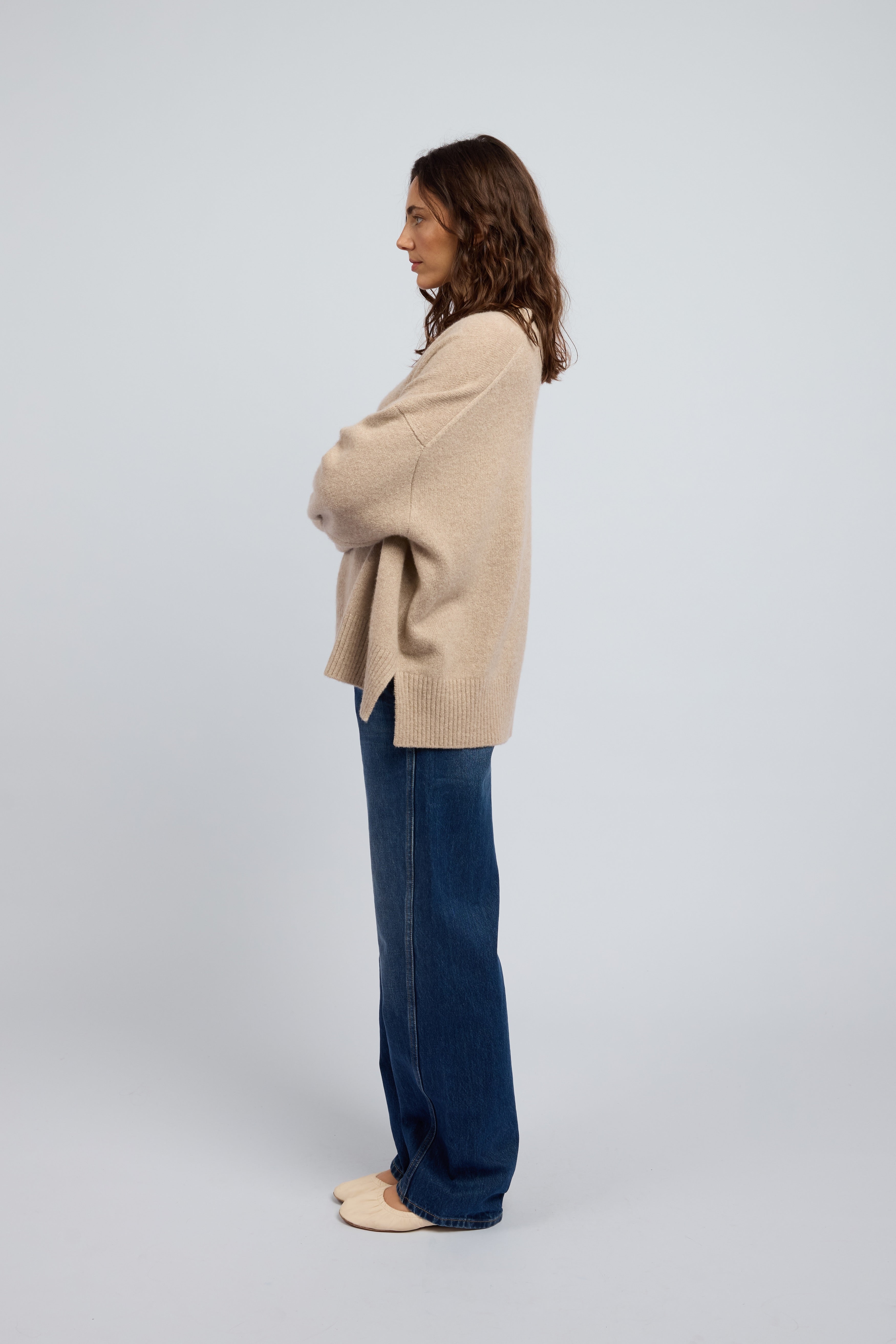 Woman wearing a slouchy beige cashmere long sleeve jumper in Oat with ribbed cuffs and neckline, paired with blue jeans and cream ballet flats